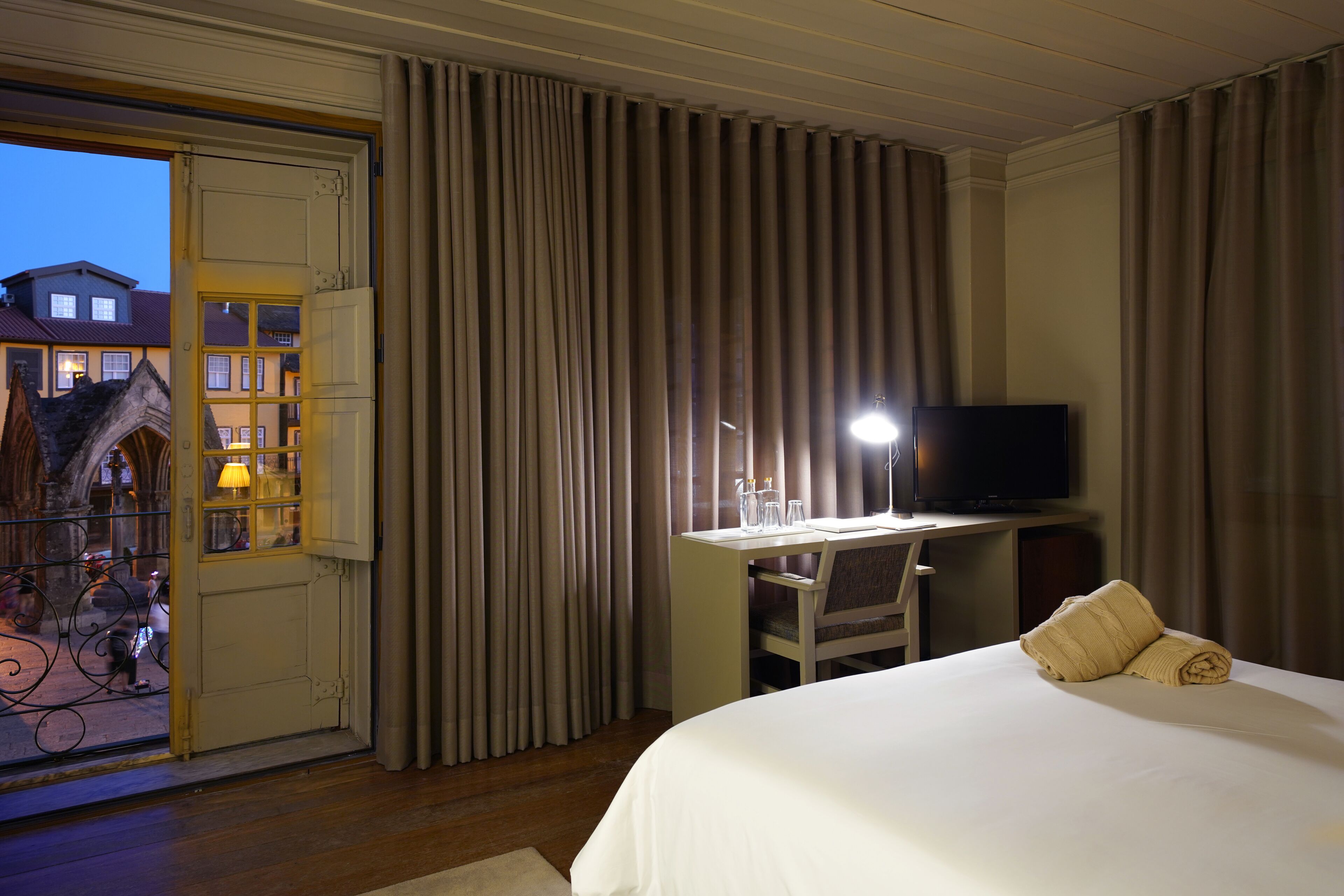 A hotel room with a bed, a desk with a television, and a view of buildings outside.