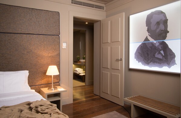 A hotel room with a bed, bedside lamp, a large portrait on the wall, and a wooden bench.