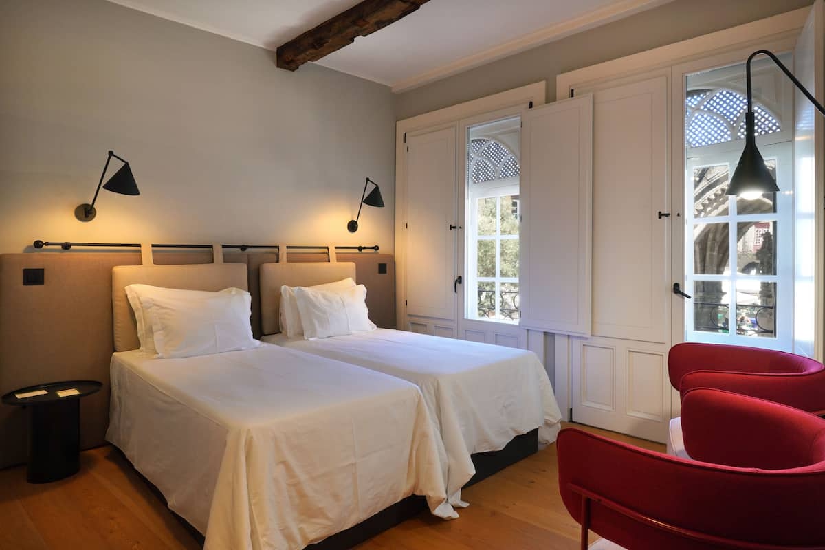 A bedroom with two beds, a red chair, and a window with a view of greenery.