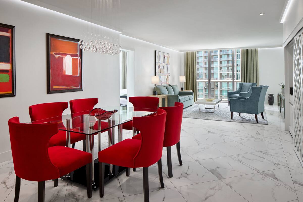 A modern living room with a glass dining table surrounded by red chairs, a sofa, and a blue armchair.