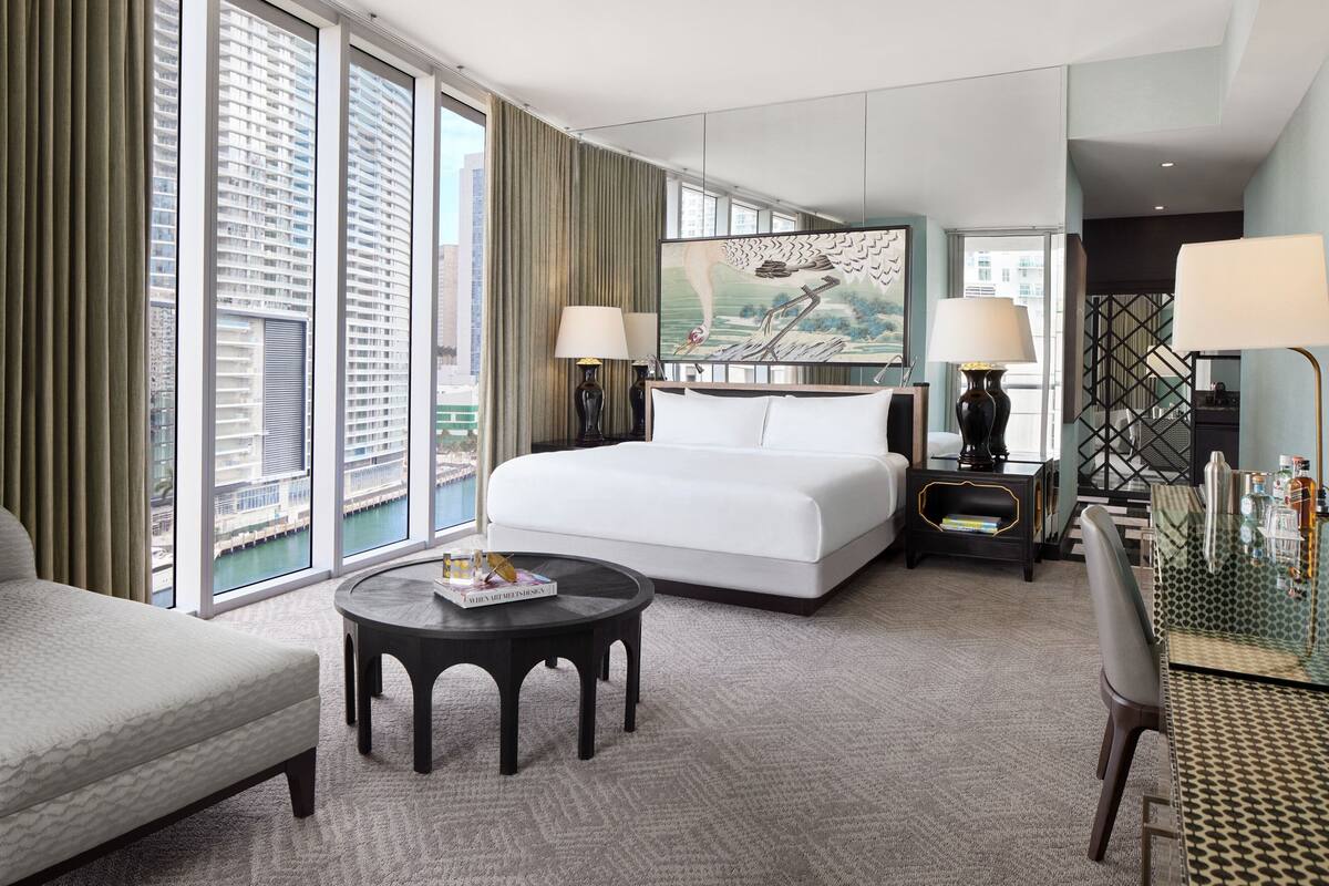 A hotel room with a large bed, a round coffee table, a desk with a lamp, and a view of the city through large windows.