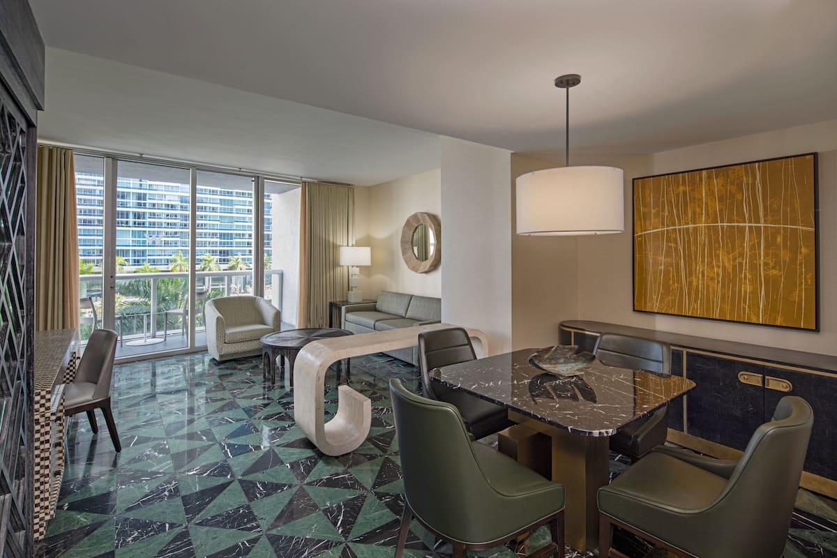 A modern hotel room with a large window, a dining area with a marble table, and a view of the cityscape.