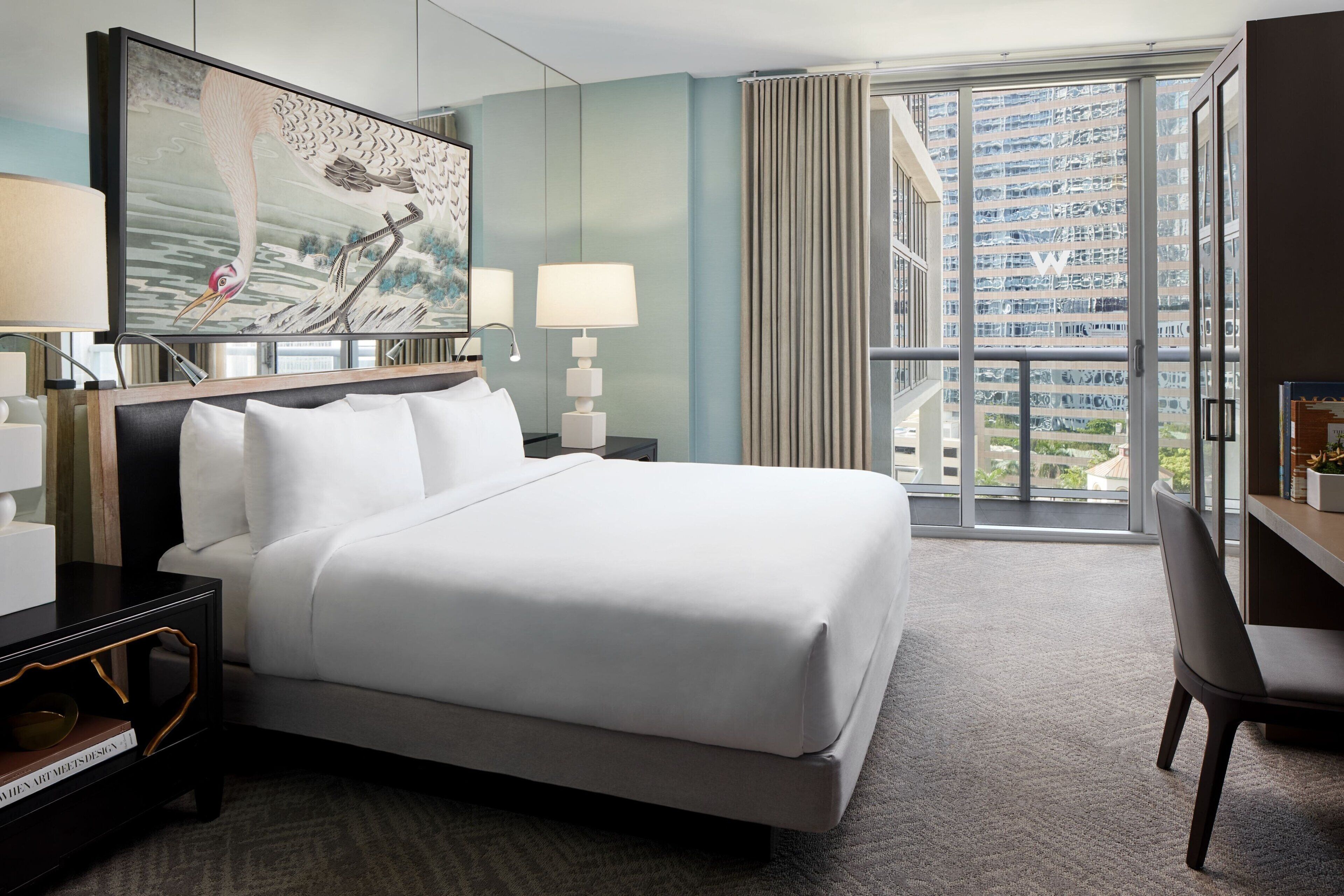 A hotel room with a large bed, a desk, a chair, and a large window with a view of a building.