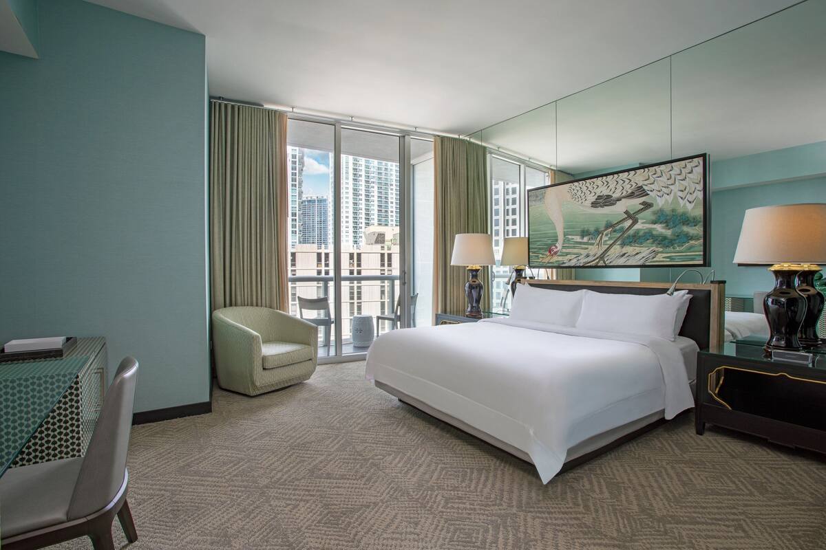 A hotel room with a large bed, a desk, a chair, and a view of tall buildings through the window.