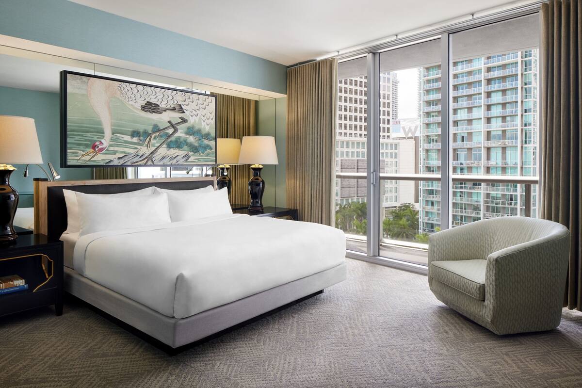 A hotel room with a large bed, two bedside tables with lamps, a chair, a large window with a city view, and a painting on the wall.