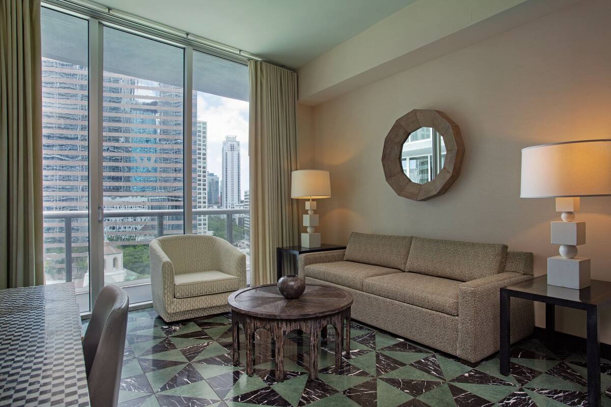 A hotel room with a view of a cityscape, a round coffee table, a sofa, a chair, and two lamps.