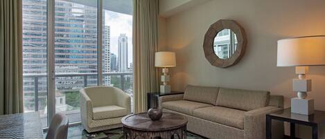 A hotel room with a view of a cityscape, a round coffee table, a sofa, a chair, and two lamps.