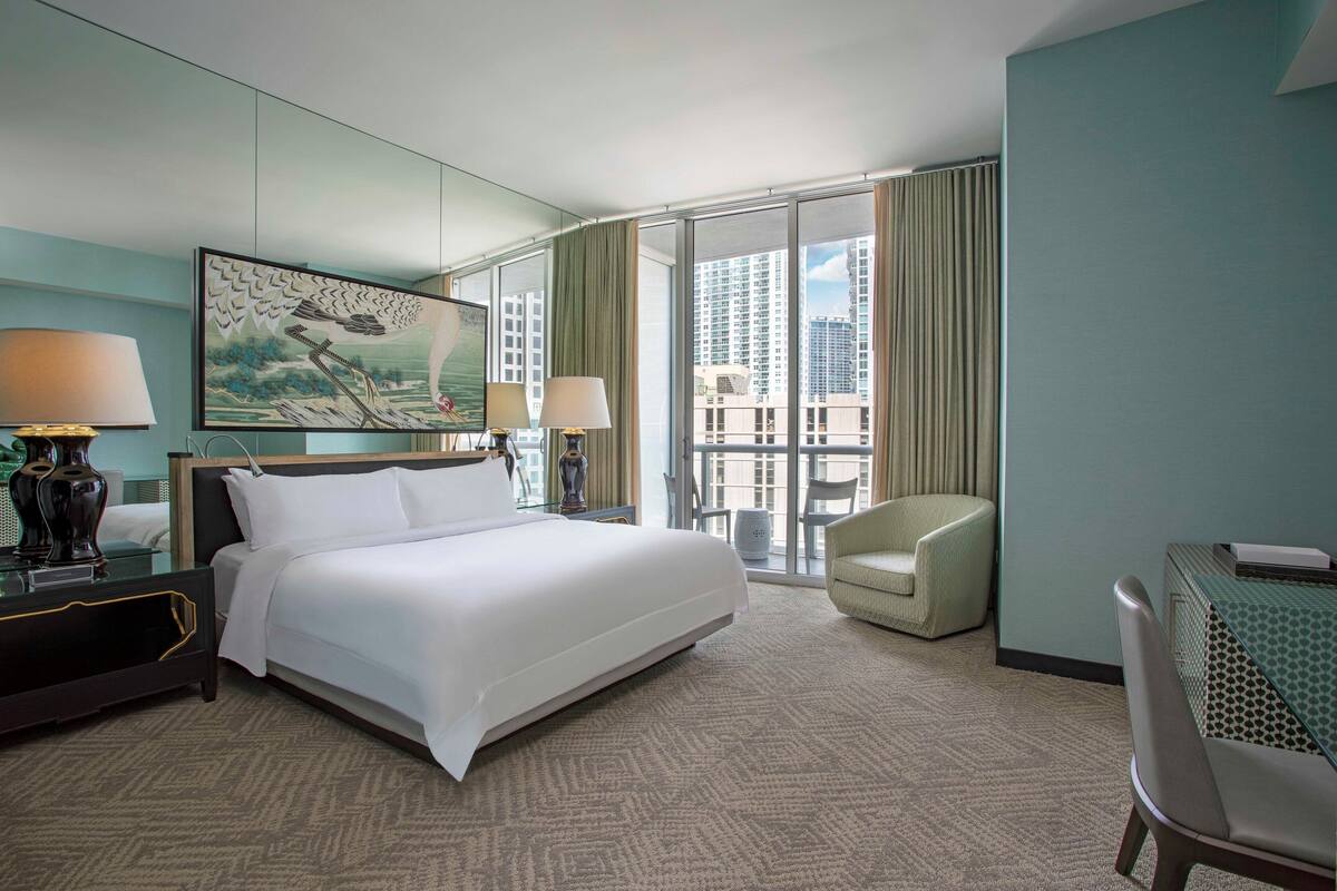 A hotel room with a large bed, two bedside tables, a desk, a chair, and a large window with a city view.