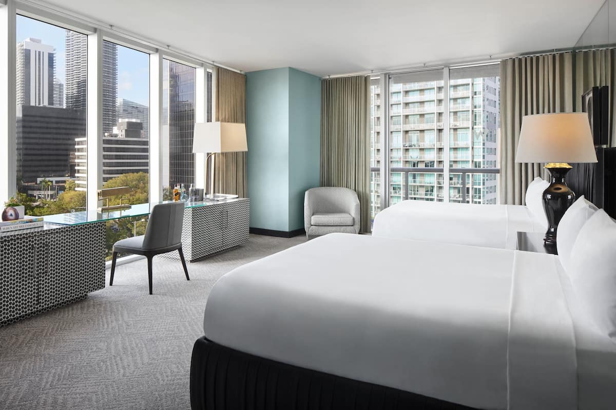 A hotel room with a large bed, a desk with a chair, a small table with a lamp, and a view of city buildings through large windows.