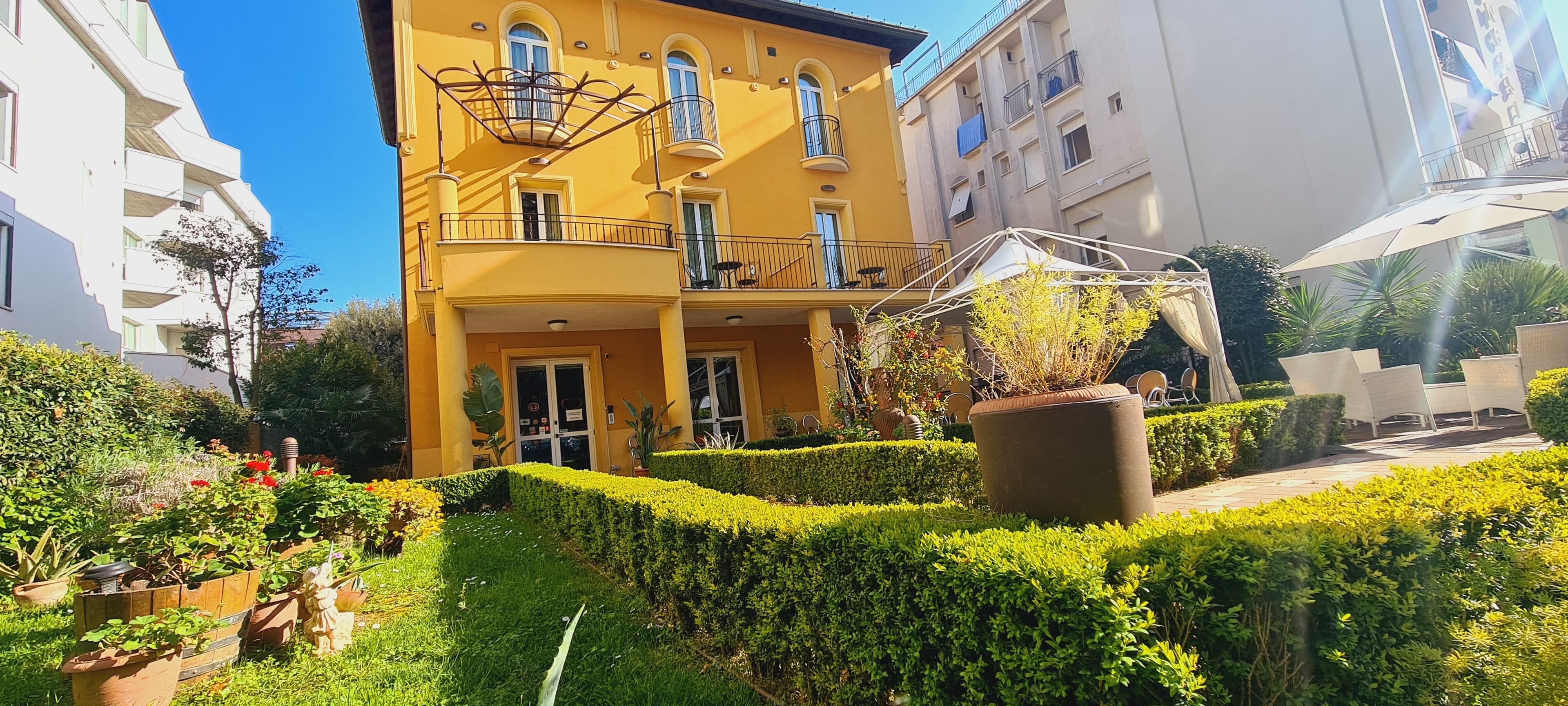 Un bâtiment jaune doté d’un balcon, entouré de haies bien entretenues et de plantes en pot.