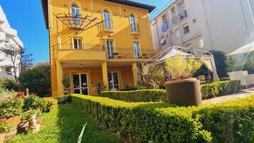 Un bâtiment jaune doté d’un balcon, entouré de haies bien entretenues et de plantes en pot.