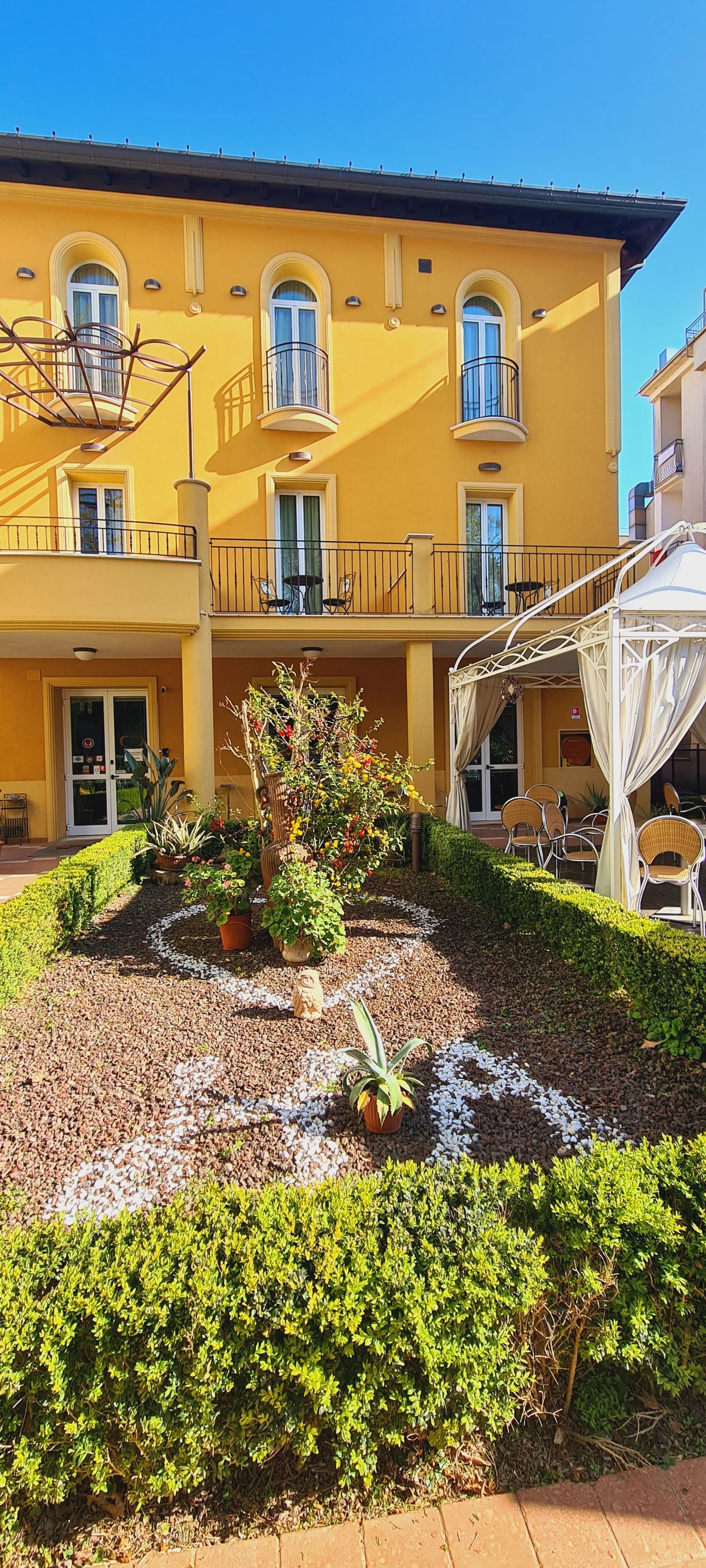 A yellow building with multiple balconies, a garden with a variety of plants, and outdoor seating.