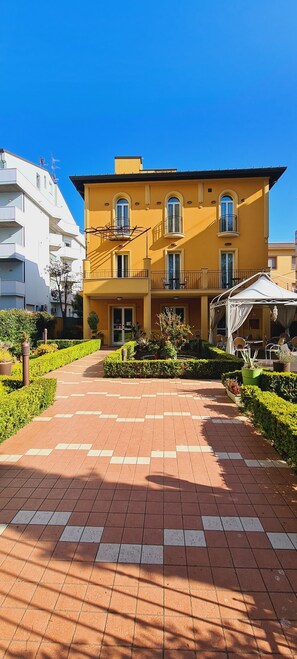 A yellow building with a paved walkway leading to a covered outdoor seating area. - Hotel Alibì (Rimini)