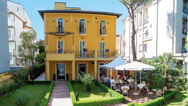 A yellow building with a covered outdoor seating area and a pathway leading to the entrance. - Hotel Alibì (Rimini)