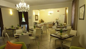 A hotel dining area with multiple tables and chairs, a chandelier, and a view of the kitchen in the background. - Hotel Alibì (Rimini)