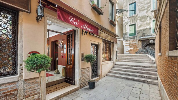 A narrow street with a building featuring a red awning and a staircase leading to an entrance. - Hotel Ca' Alvise (Venice)