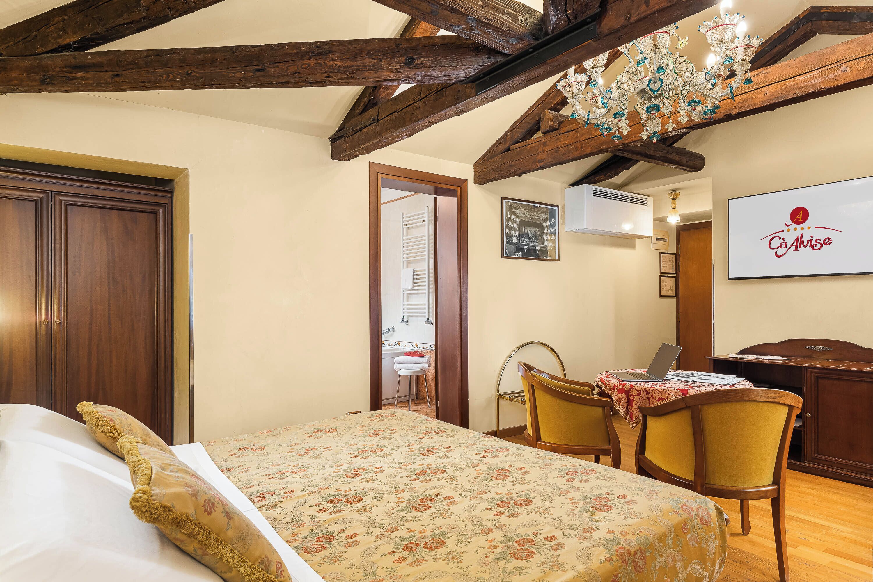 A bedroom with a bed, wooden ceiling beams, a chandelier, a desk with a chair, and a wall-mounted TV.