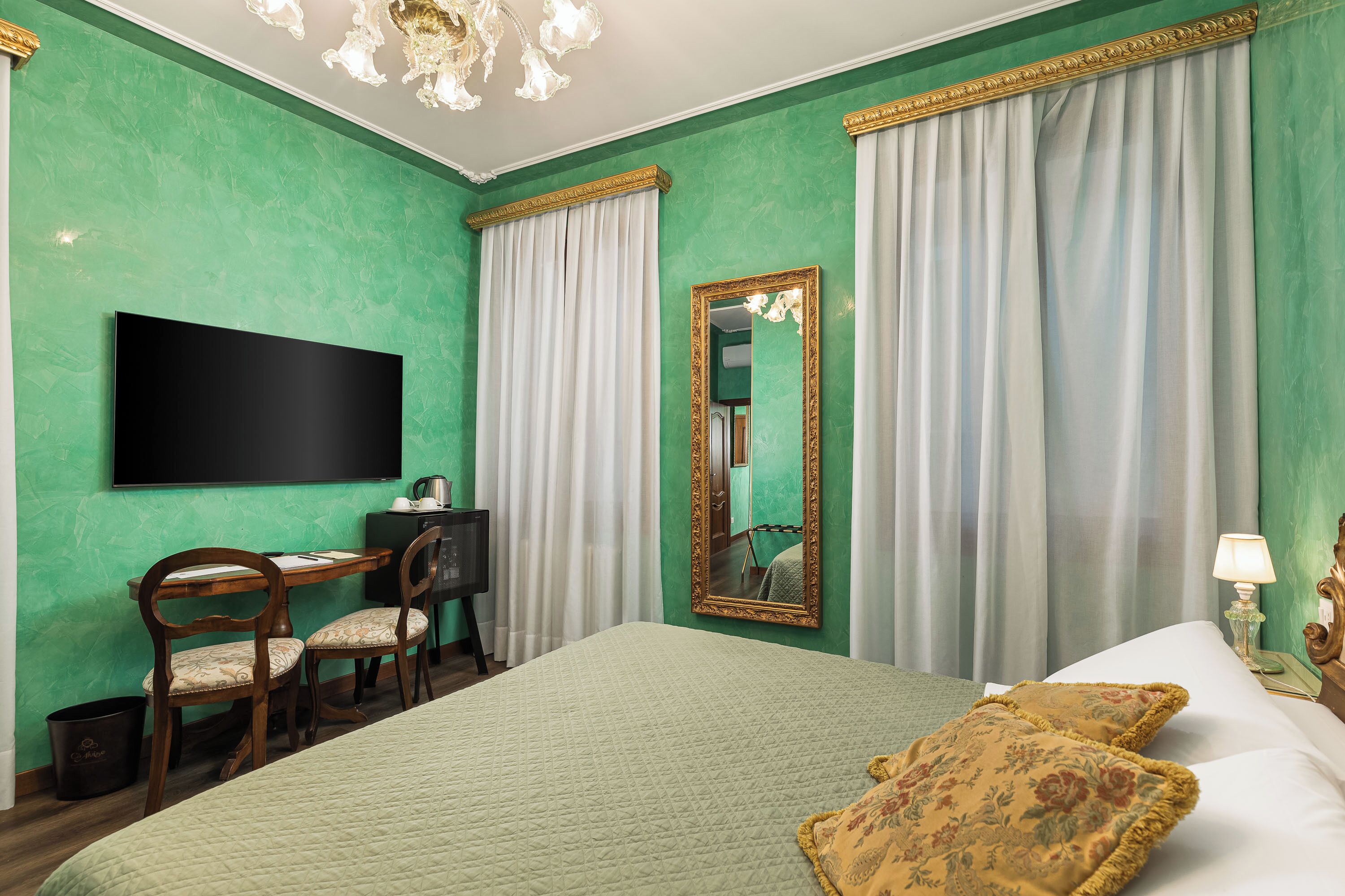 A room with a bed, a desk with chairs, a television, a mirror, and curtains.