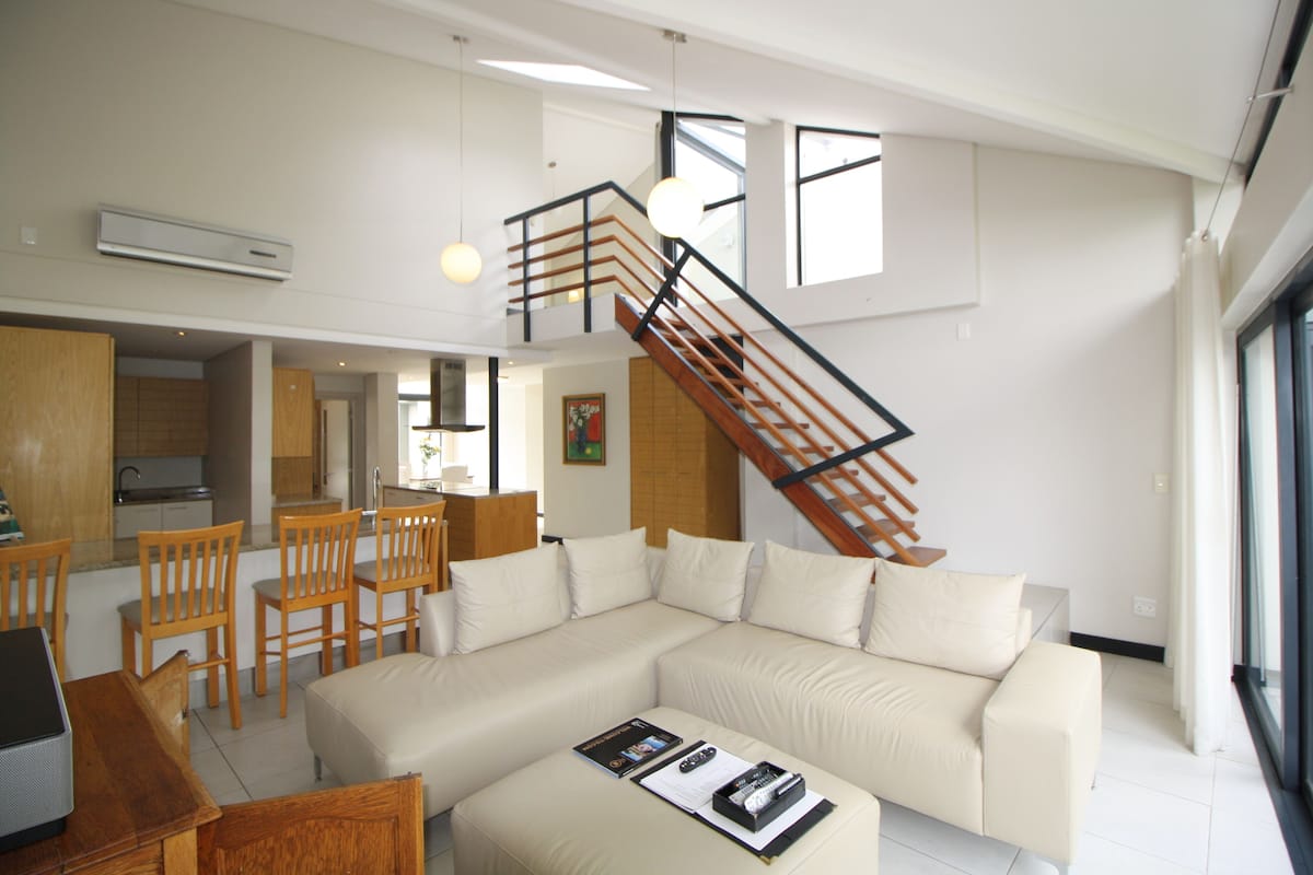 Uma sala de estar moderna com um amplo sofá em formato de L, cadeiras de madeira, uma mesa de centro e uma escada que leva a um andar superior.