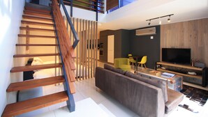 A modern living space with a wooden staircase, a grey sofa, a coffee table, a TV, and a dining area with yellow chairs. - The Glen Apartments (Cape Town)