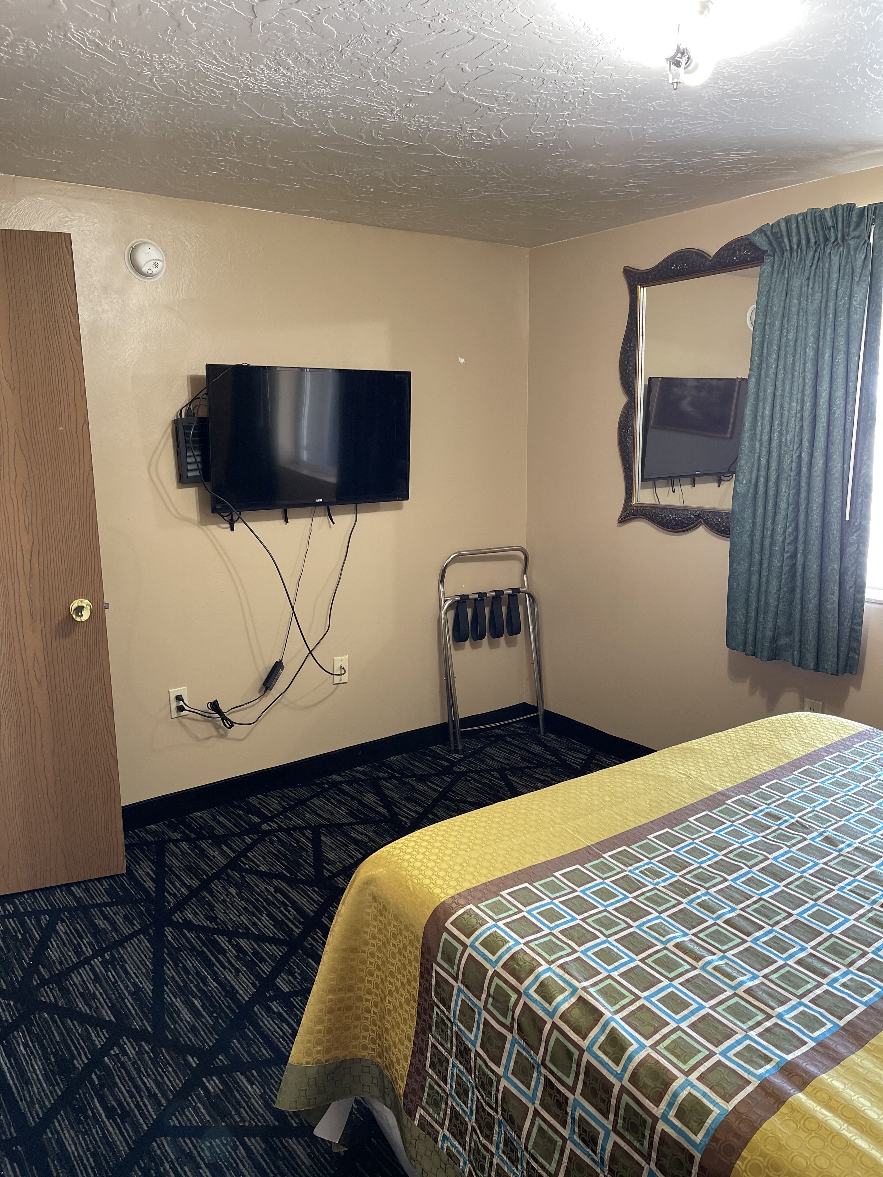 A hotel room with a bed, a television mounted on the wall, a mirror, and a shoe rack.