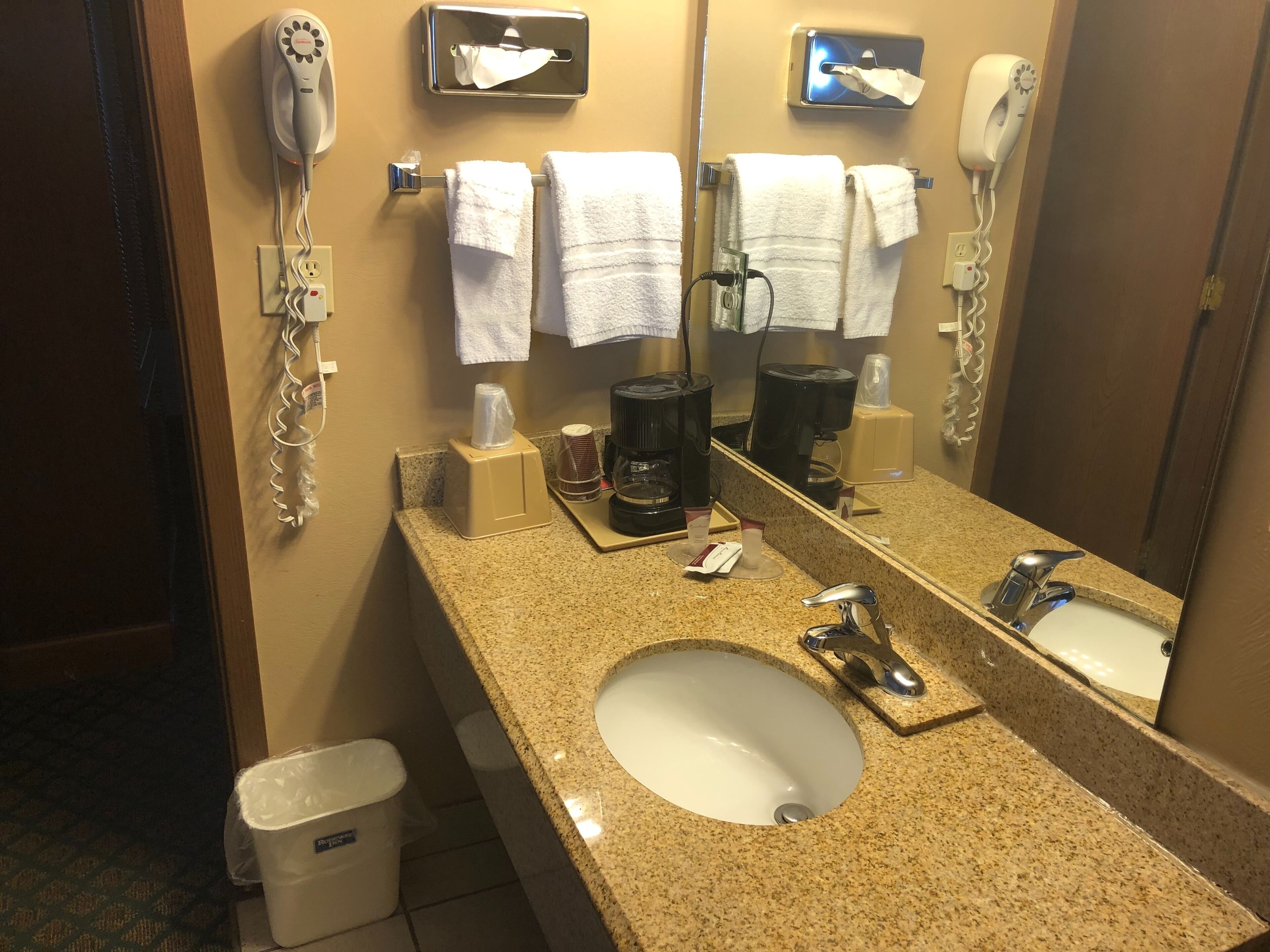 A hotel bathroom with a granite countertop, a sink, a mirror, a hairdryer, a towel dispenser, and a coffee maker.