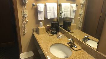 A hotel bathroom with a granite countertop, a sink, a mirror, a hairdryer, a towel dispenser, and a coffee maker.