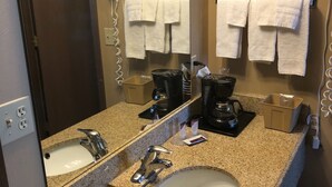 A hotel bathroom with a granite countertop, two sinks, a large mirror, a hairdryer, a coffee maker, and towels hanging on hooks. - Riverton Inn & Suites (Riverton)