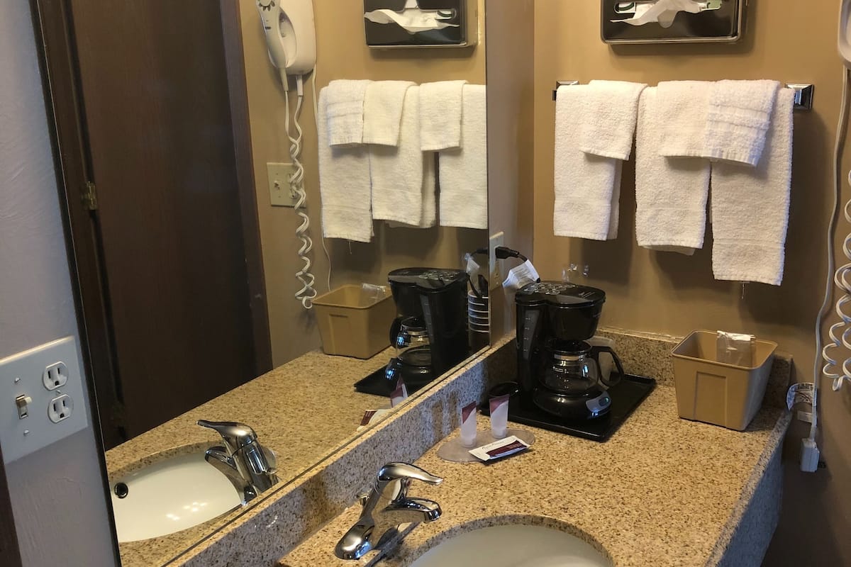 Casa de banho de hotel com bancada de granito, duas pias, um espelho grande, secador de cabelo, máquina de café e toalhas penduradas em ganchos.