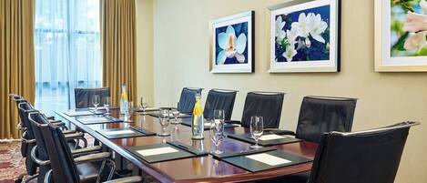 A conference room with a long table, black leather chairs, framed floral artwork on the wall, and a window with curtains.