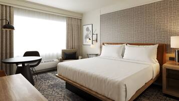 A hotel room with a large bed, a small table, a chair, a desk, and a lamp.