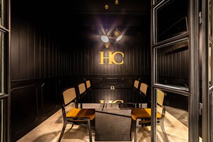 Een vergaderruimte met een glazen tafel, houten stoelen en een zwarte muur waarop de letters 'HC' verlicht zijn.