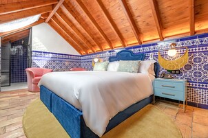 Een slaapkamer met een houten plafond, een bed met een blauw hoofdeinde, een nachtkastje en een stoel.