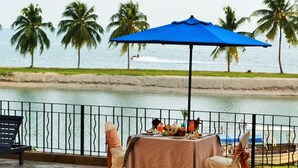 An outdoor dining area with a table set for two, overlooking a body of water with palm trees in the background. - Corus Paradise Resort Port Dickson (Port Dickson)