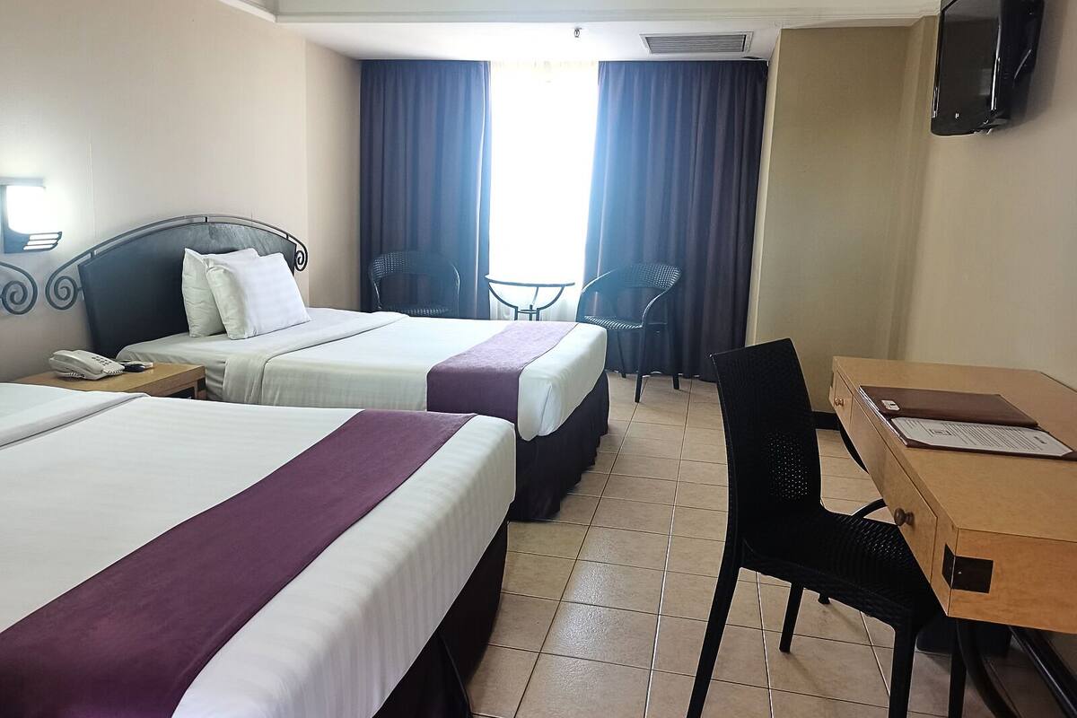 A hotel room with two beds, a desk, a chair, a TV, and a window with curtains.