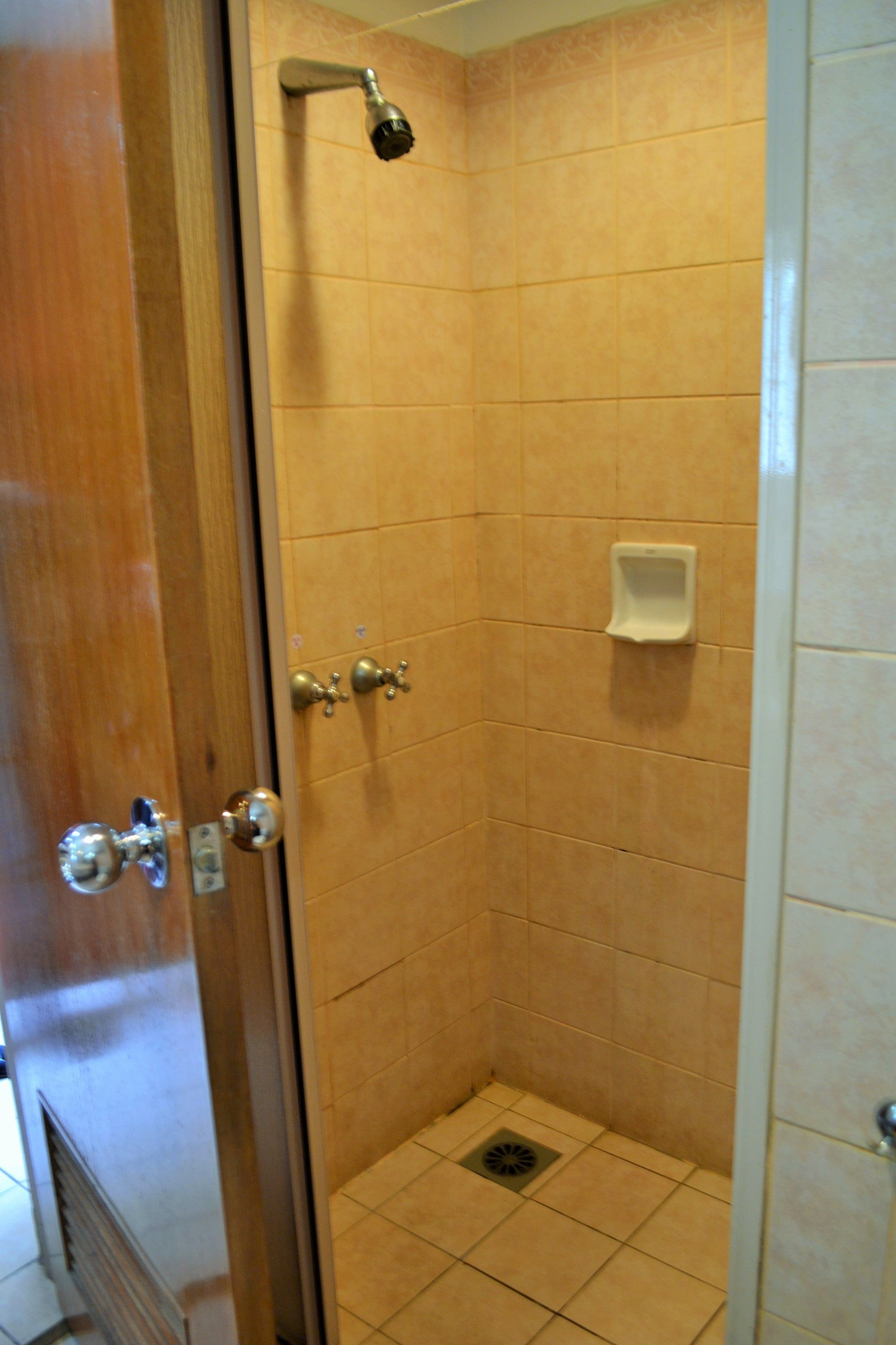 A small, enclosed shower area with tiled walls and floor, a handheld showerhead, a soap dispenser, and a door with a handle and lock.