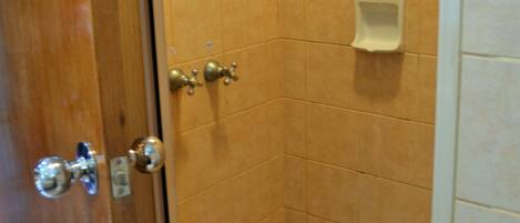 A small, enclosed shower area with tiled walls and floor, a handheld showerhead, a soap dispenser, and a door with a handle and lock.