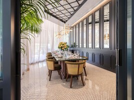 A dining area with a long table set for four, surrounded by wicker chairs, with a large window and a plant on the left.