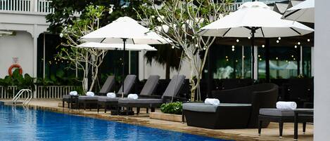 A poolside area with lounge chairs and umbrellas, a pool, and a building with balconies in the background.