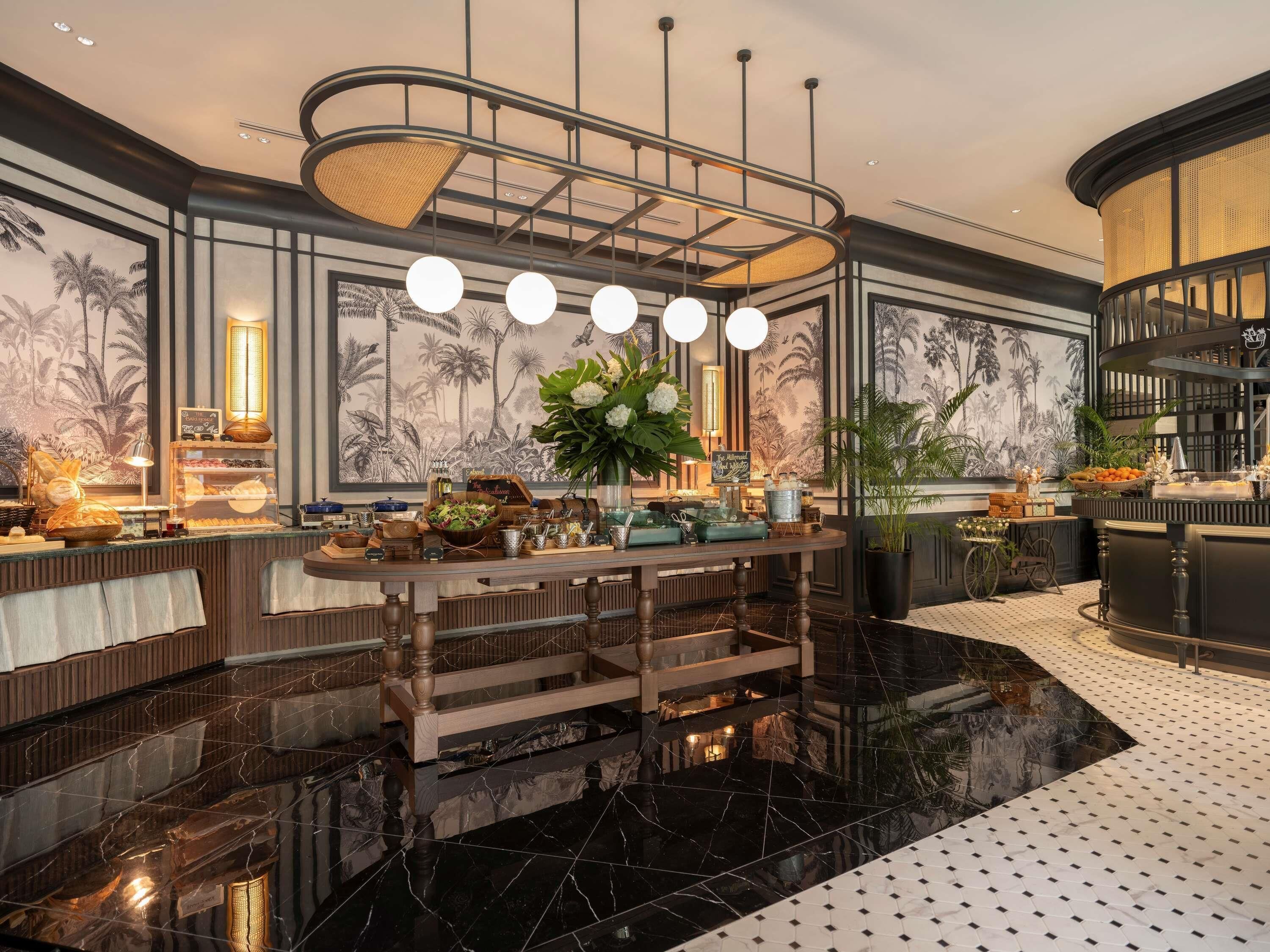 A luxurious hotel lobby with a marble floor, a central bar area with a wooden counter, and tropical-themed wall art.