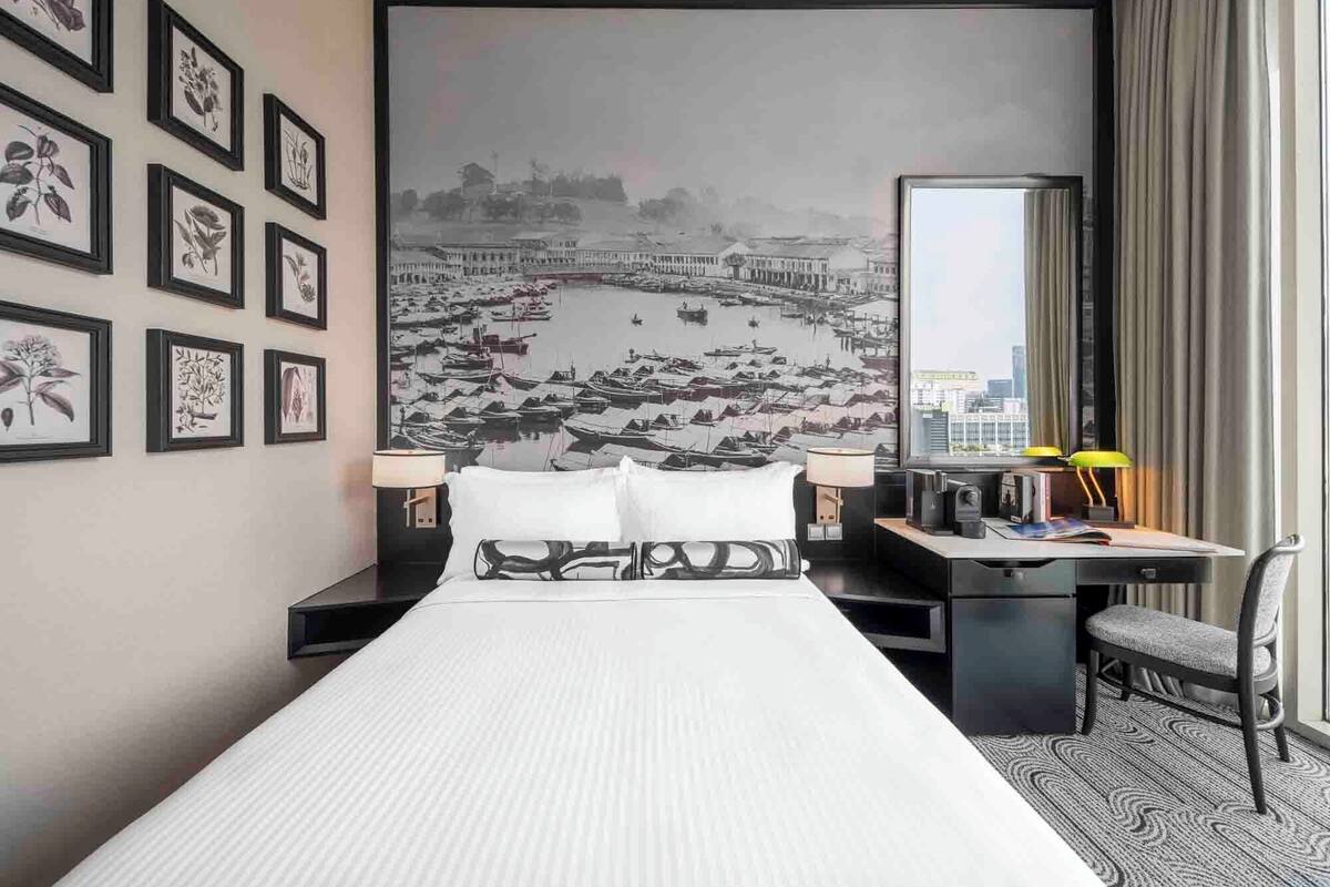 A hotel room with a large bed, a desk with a chair, a desk lamp, and a large window with curtains.