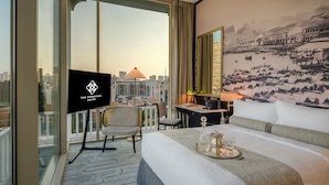Habitación de hotel con una cama grande, un escritorio, una silla, un televisor y vistas a la ciudad a través de amplias ventanas.