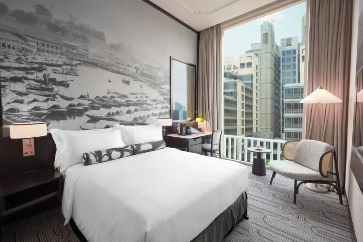 A hotel room with a large bed, a desk, a chair, a balcony with a view of buildings, and a wall with a mural.