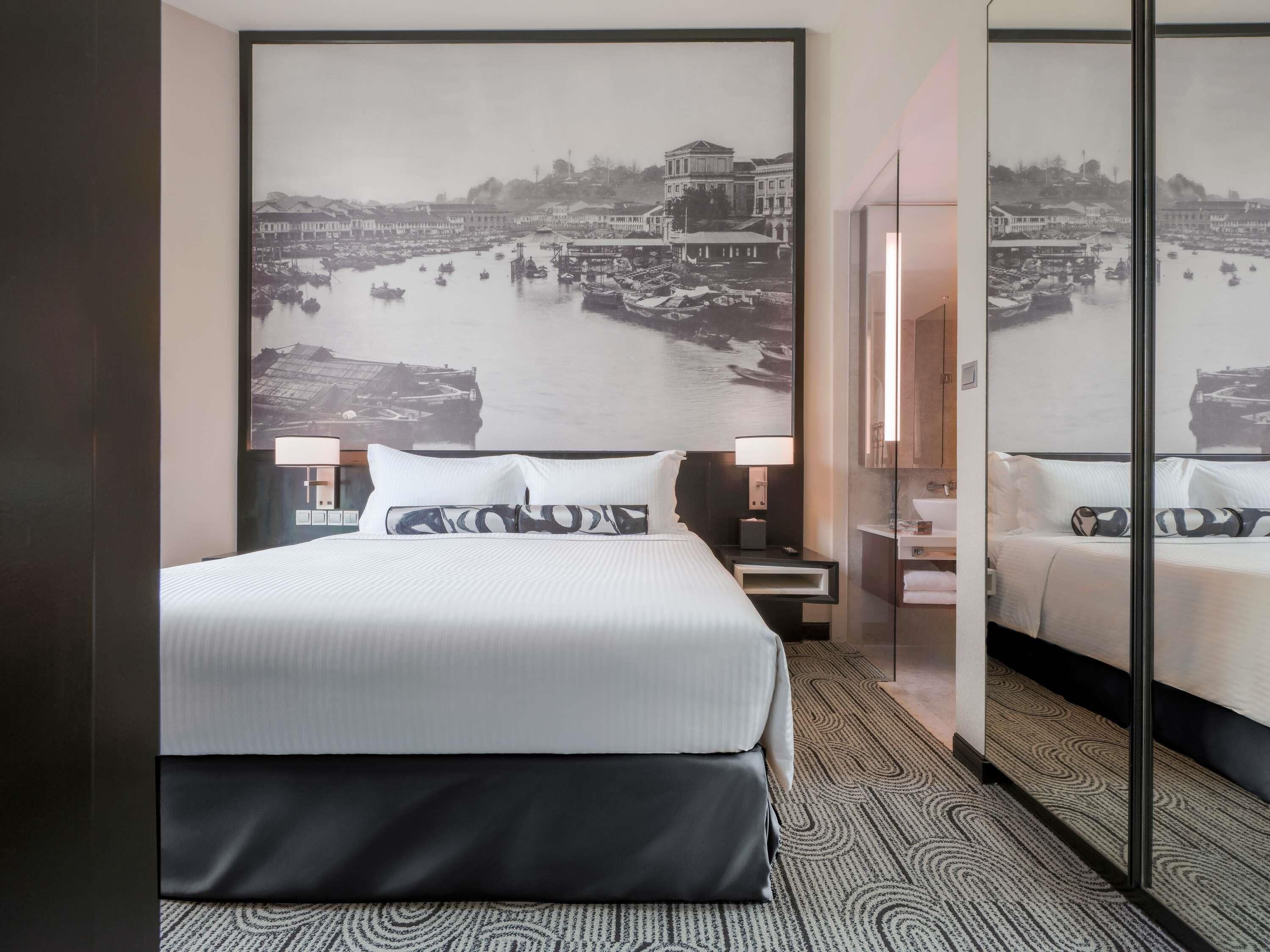 Une chambre d’hôtel avec un grand lit, une table de chevet, une commode, un miroir et un mur orné d’une photographie en noir et blanc.