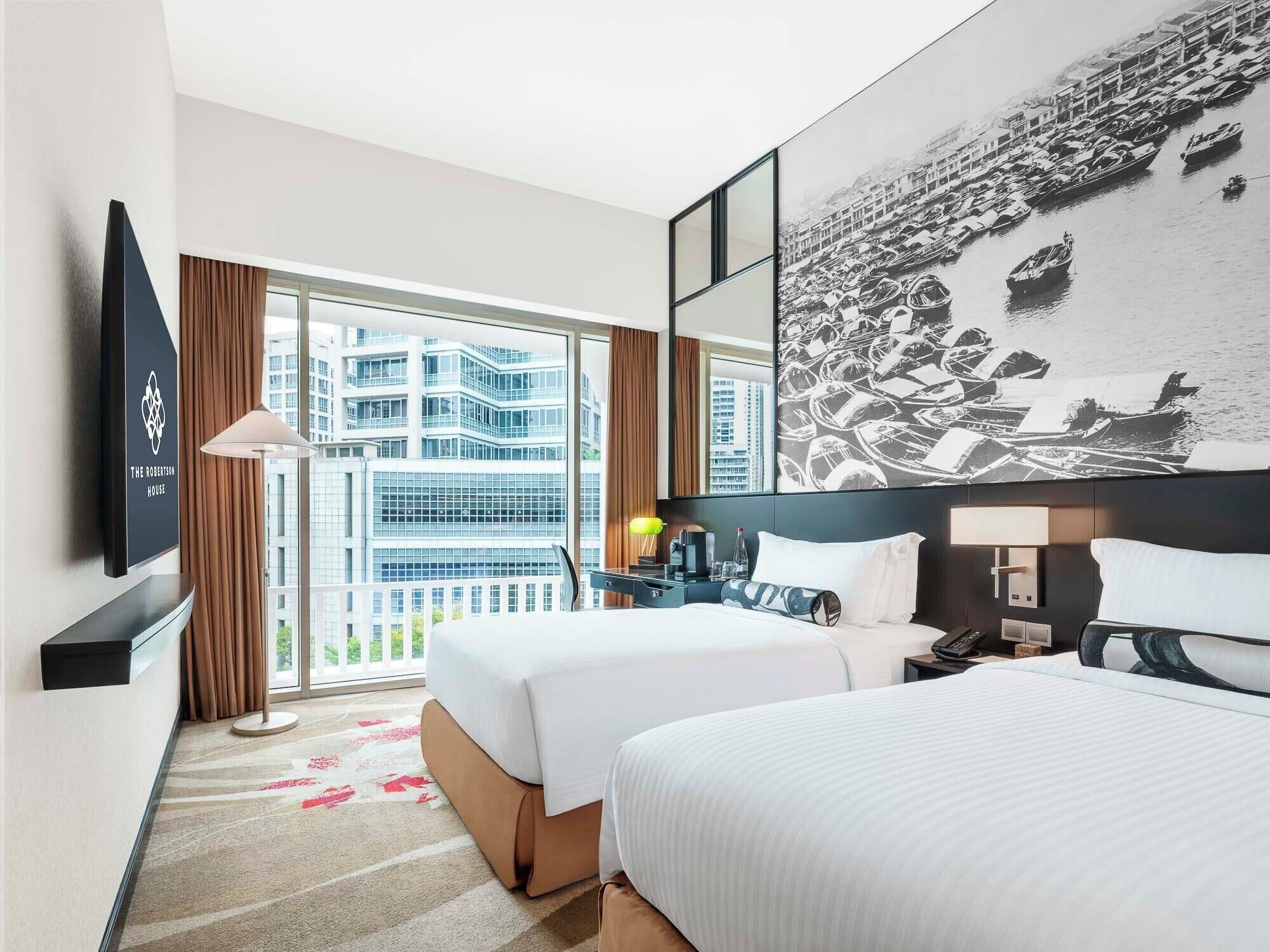 Habitación de hotel con dos camas, un ventanal con vistas a edificios, un televisor de pantalla plana y un cuadro de barcos.