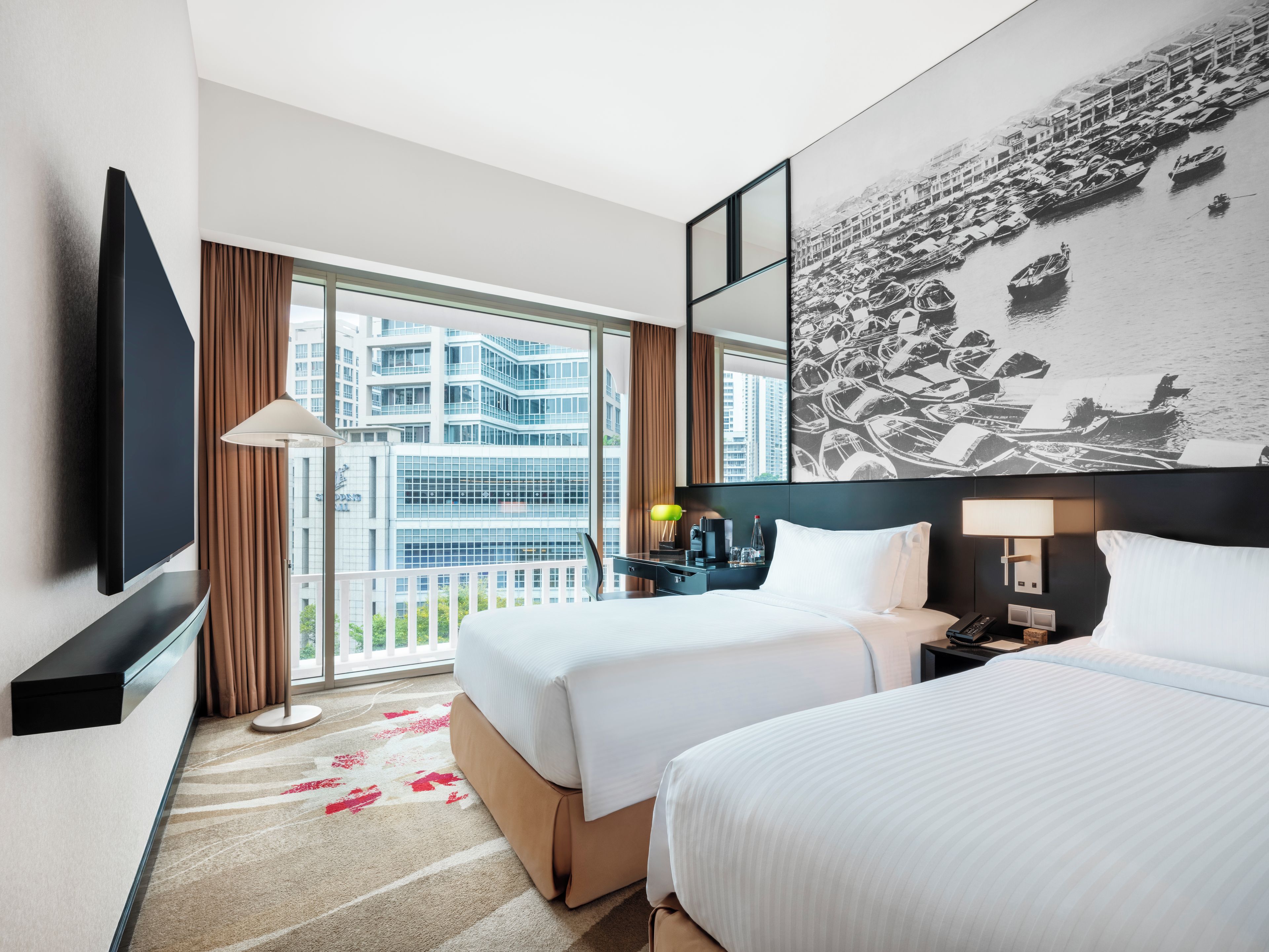A hotel room with two beds, a large window with a city view, a flat-screen TV, and a wall-mounted artwork.