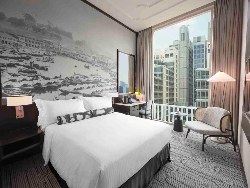 Habitación de hotel con una cama grande, un escritorio, una silla, una mesita, una lámpara y vistas a edificios a través del ventanal.