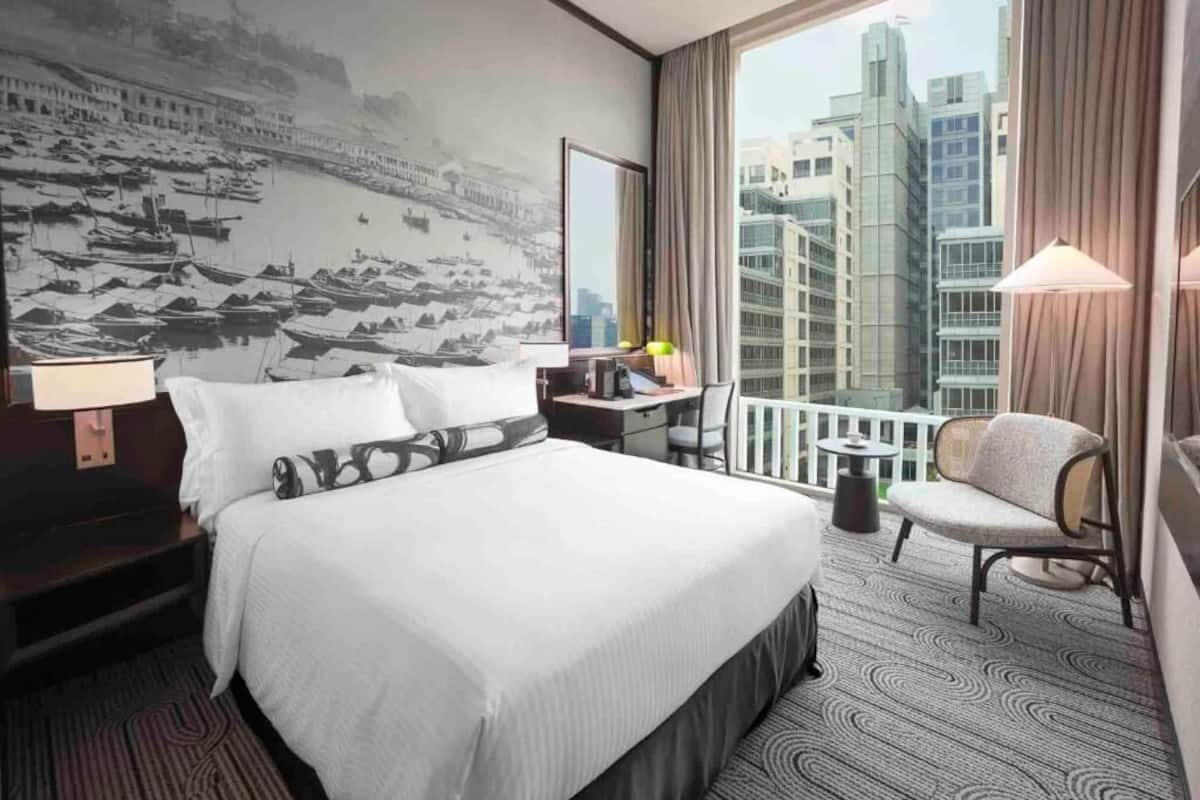 Habitación de hotel con una cama grande, un escritorio, una silla, una mesita, una lámpara y vistas a edificios a través del ventanal.