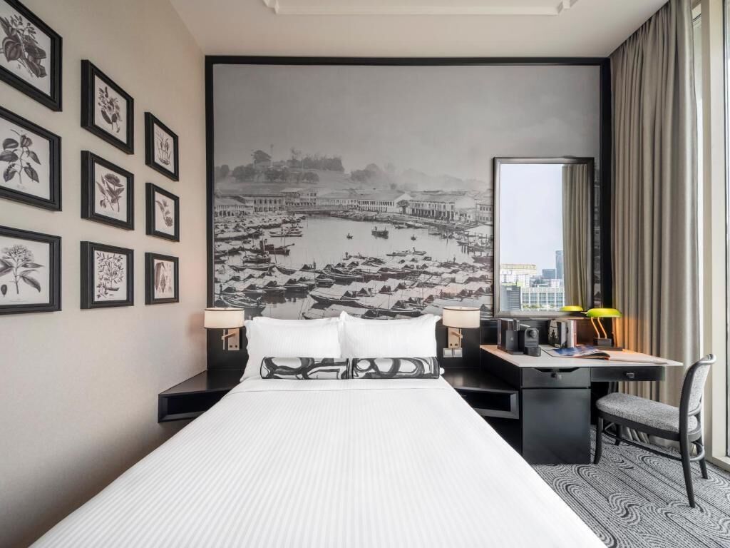 Una habitación de hotel con cama, escritorio, silla y un amplio ventanal con un mural de paisaje urbano en la pared.
