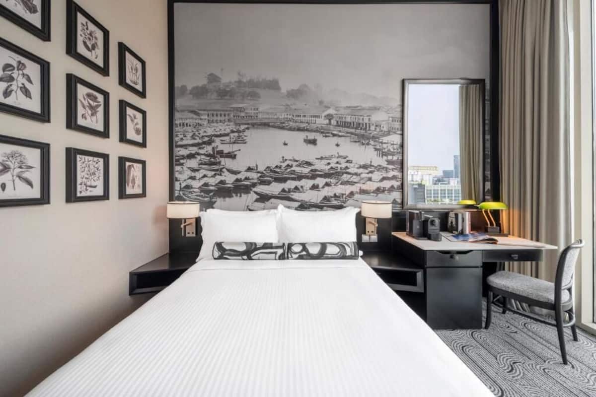 Una habitación de hotel con cama, escritorio, silla y un amplio ventanal con un mural de paisaje urbano en la pared.